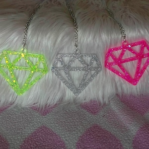 May include: Three diamond-shaped pendants hanging from silver-toned chains. The left pendant is neon green with glitter, the center pendant is silver with glitter, and the right pendant is bright pink with glitter, all featuring a geometric frame.