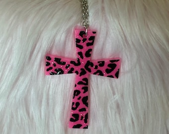 May include: A cross pendant necklace featuring a bright pink base with a black leopard print pattern and subtle glitter accents. The cross is suspended from a silver-toned metal chain that attaches to the top of the pendant through a small loop.