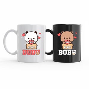 May include: A pair of ceramic mugs, one white and one black, with matching cartoon bear and panda designs. The white mug features a panda holding a heart, with the text "He is my Dudu". The black mug has a bear with the text "She is my Bubu".
