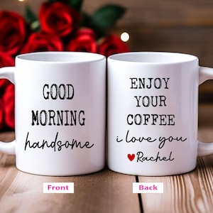 May include: Two white ceramic coffee mugs. One mug reads "GOOD MORNING handsome" in black script. The other mug reads "ENJOY YOUR COFFEE i love you ❤️ Rachel" in black script with a red heart. Red roses are in the background.