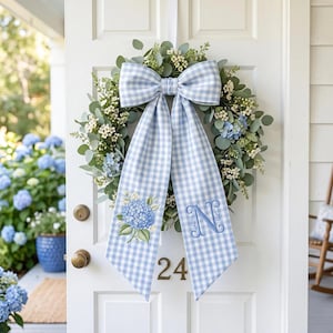 May include: A circular wreath featuring green eucalyptus leaves, small white flowers, and blue hydrangea clusters. A large light blue and white gingham bow hangs from the top, with one tail embroidered with a blue hydrangea and the other with a blue letter N.