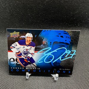 Könnte beinhalten: Eine Hockey-Sammelkarte mit Connor McDavid von den Oilers. Die Karte hat ein blaues und weißes Farbschema, wobei der Spieler in Aktion ist. Die Karte enthält auch den Text "COMPOSITIONS" und "97".