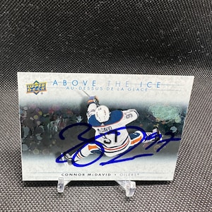 以下が含まれることがあります： OilersのConnor McDavidをフィーチャーしたホッケーカード。「ABOVE THE ICE」と「AU-DESSUS DE LA GLACE」の文字が入っています。McDavidのサインは青インクでカード全体に書かれています。
