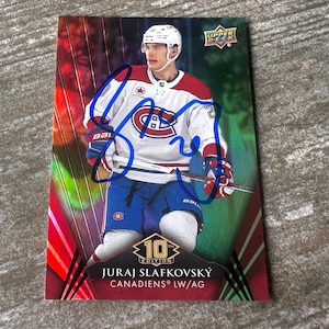 Könnte beinhalten: Eine rechteckige Upper Deck 10th Edition Eishockey-Sammelkarte mit Juraj Slafkovský in einem weiß-rot-blauen Trikot der Montreal Canadiens. Die Karte hat einen leuchtend roten und grünen Hintergrund mit einem Autogramm in blauer Tinte in der Mitte.