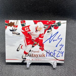 Könnte beinhalten: Eishockey-Sammelkarte mit Pavel Datsyuk im roten Trikot der Detroit Red Wings. Die Karte hat ein Autogramm und den Text "HOF 24". Der Hintergrund zeigt andere Spieler und die Eisfläche.