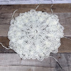 Puede incluir: Un tapete blanco y circular con un intrincado diseño de encaje. El tapete presenta un patrón floral con delicados detalles calados. Varios hilos sueltos se extienden desde el borde. El tapete está colocado sobre una superficie de madera.