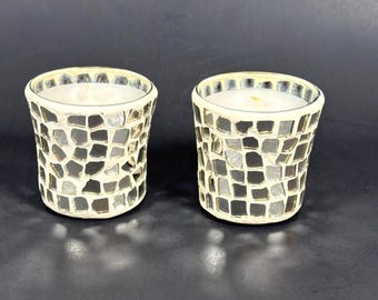 Puede incluir: Dos candelabros de cristal de mosaico con velas blancas. Los candelabros están decorados con pequeñas baldosas de espejo de forma irregular en plata y crema. Los candelabros cilíndricos tienen un borde grueso de color crema.