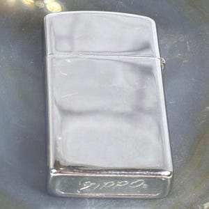 May include: A shiny, chrome-colored Zippo lighter. The rectangular lighter has rounded edges and a hinged lid. The brand name "Zippo" is embossed on the bottom. The surface reflects the surrounding environment.