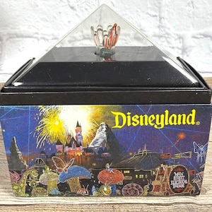 May include: A Disneyland souvenir box with a clear, pyramid-shaped top. The box features a colorful illustration of fireworks, a castle, and other attractions. The Disneyland logo is prominently displayed in yellow.