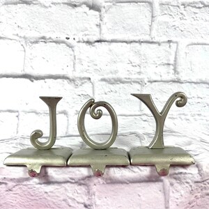 Op de afbeelding: Zilveren metalen kerstsokkenhouders met het woord "JOY". Elke letter is bevestigd aan een rechthoekige basis met een kleine haak. De achtergrond is een witte bakstenen muur.