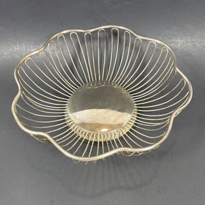 Könnte beinhalten: Silberfarbener Draht-Obstkorb mit gewelltem Rand. Der Korb hat einen kreisförmigen Boden und ein radiales Drahtdesign, das ein offenes Aussehen erzeugt. Der Korb ist für Obst oder andere Gegenstände konzipiert.