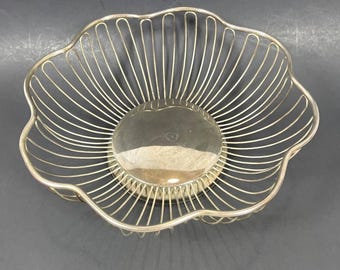 Puede incluir: Cesta de frutas de alambre plateado con borde festoneado. La cesta tiene una base circular y un diseño de alambre radial, creando una apariencia calada. La cesta está diseñada para contener frutas u otros artículos.