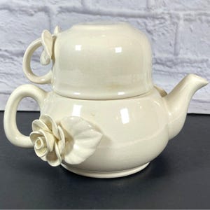 May include: Cream-colored ceramic teapot with a matching teacup. The teapot features a rose and leaf design on the side and handle. The teacup sits atop the teapot, creating a stacked appearance. The teapot has a spout for pouring.