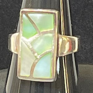 May include: A silver ring with a rectangular face. The face is inlaid with iridescent, light green and white pieces, creating a mosaic pattern. The ring is set on a black background.