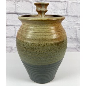 May include: A ceramic jar with a lid featuring a tapered body and a flared rim. The exterior displays a gradient of colors, transitioning from a dark grey base with horizontal ridges to a speckled olive green middle and a warm tan top section.