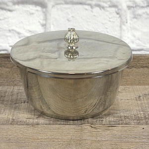 May include: A silver-colored, round container with a lid. The lid has a decorative knob on top. The container sits on a wooden surface. The background is a white brick wall.
