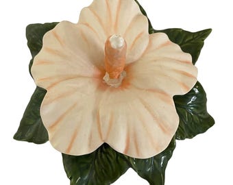 Puede incluir: Una figura de cerámica con forma de flor de hibisco que presenta cinco pétalos de color melocotón pálido con vetas anaranjadas sutiles y un estambre central. La flor descansa sobre una base de hojas verde oscuro, brillantes y texturizadas.
