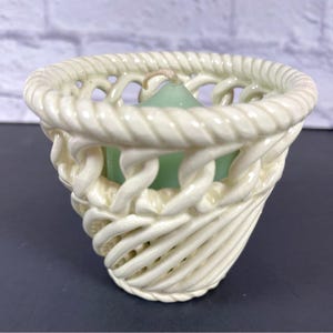May include: Cream-colored ceramic candle holder with a woven basket design. The candle holder has a green candle inside. The basket weave is detailed and the candle holder is on a dark surface.