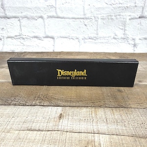 Może przedstawiać: Czarny prostokątny pojemnik z logo Disneylandu w złotych literach. Pojemnik stoi na drewnianej powierzchni z białą ceglaną ścianą w tle. Logo Disneylandu zawiera słowa "Southern California."