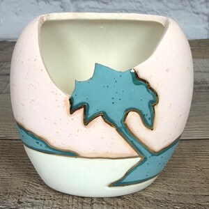 May include: A ceramic, egg-shaped planter with a light pink exterior and a turquoise leaf design. The planter has a white base and interior, and the leaf design is outlined in gold. The planter is suitable for small plants or decorative use.