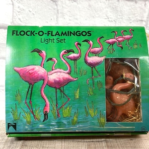 May include: A vintage "Flock-O-Flamingos" light set in a green box. The box features illustrations of pink flamingos in a marsh setting. The light set includes several flamingo-shaped lights. The text "Light Set" is printed on the box.