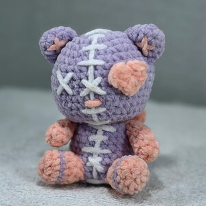 May include: A plush crochet bear toy in lavender and peach yarn. The bear features a white stitched seam running down the centre of its head and torso, a peach heart-shaped patch over one eye, and white X-shaped stitches for the other eye.