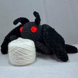 May include: A black crocheted moth plush toy with two large, round, glossy red eyes and prominent antennae. The moth is positioned behind a small, round ball of white yarn, showcasing a textured, soft-looking yarn construction throughout.