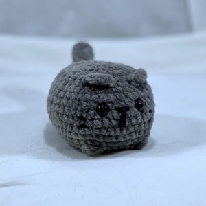 May include: A handmade gray crochet cat with black safety eyes and a small tail. The plush cat is round and has small, pointed ears. The cat is made of a soft, textured yarn.