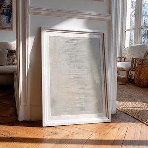 May include: A framed abstract art print with a white border leans against a white wall. The artwork features horizontal lines in shades of gray and beige. The room has hardwood floors and a view of a building through a window.