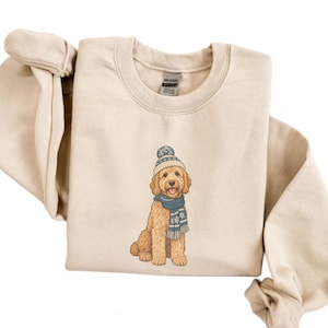 May include: A beige crewneck sweatshirt featuring a graphic of a golden-coloured dog wearing a blue and white knit beanie and a matching blue and white scarf with snowflake patterns. The Gildan brand tag is visible at the inner neckline.