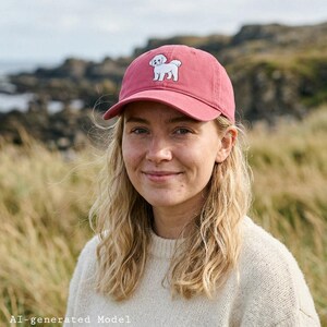 May include: A dusty rose baseball cap featuring an embroidered white Bichon Frise dog on the front panel. The cap has a curved brim, a structured crown, and a matching fabric strap with a metal buckle closure at the back for an adjustable fit.