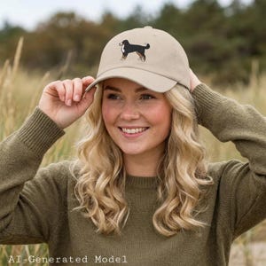 May include: A beige baseball cap featuring an embroidered graphic of a black, white, and brown dog standing in profile. The cap has a curved brim and a structured crown, showcasing a detailed canine illustration centered on the front panel.