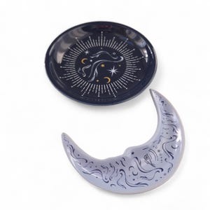 May include: Two ceramic trinket dishes. One is a dark blue, round dish with a celestial design in white and gold. The other is a light blue crescent moon-shaped dish with a detailed celestial pattern in dark blue. Both are on a white background.