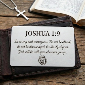 Può includere: Una carta portafoglio argentata con il testo "JOSHUA 1:9" e un versetto biblico. La carta è in un portafoglio in pelle marrone, con una collana con croce argentata e un libro aperto sullo sfondo.