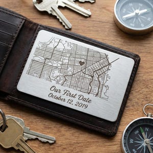May include: A brown leather wallet with a silver card featuring a map and the text "Our First Date October 12, 2019." Keys and a compass are on the wooden surface.