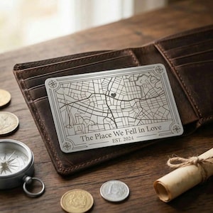 Peut inclure: Une carte en métal argenté avec un plan de ville, un symbole de cœur et le texte "The Place We Fell in Love EST. 2024" est posée dans un portefeuille en cuir marron. Une boussole, des pièces et un parchemin roulé sont également présents.