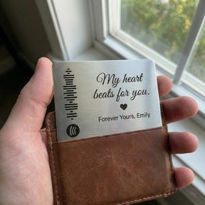 Peut inclure: Une carte en métal argenté avec le texte "My heart beats for you" et "Forever Yours, Emily" est tenue dans un portefeuille en cuir marron. La carte présente un motif d'onde sonore sur le côté gauche et un symbole de cœur.