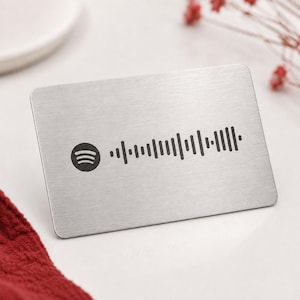 Puede incluir: Una tarjeta rectangular de metal plateado cepillado con un logotipo de Spotify negro en el lado izquierdo, seguido de un código de onda sonora escaneable de Spotify en negro. La tarjeta tiene esquinas redondeadas y acabado metálico.