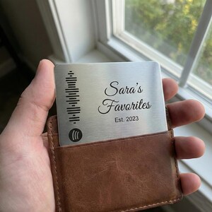 Puede incluir: Una tarjeta de metal plateado con un código de Spotify y el texto "Sara's Favorites Est. 2023" se sostiene en un tarjetero de cuero marrón. La tarjeta tiene un diseño elegante y minimalista.