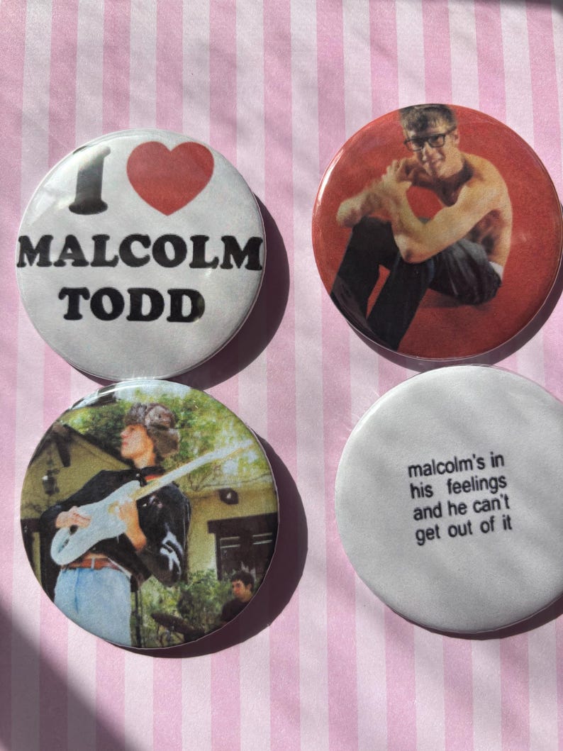 Op de afbeelding: Vier ronde buttons met afbeeldingen en tekst. E&eacute;n button zegt "I ❤️ MALCOLM TODD" met een rood hart. Een andere toont een man met een bril. Een derde toont een man die gitaar speelt. De laatste zegt "malcolm's in his feelings and he can't get out of it."