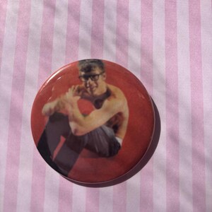 Op de afbeelding: Een ronde, rode button met een foto van een persoon met een bril die zit. De button heeft een glanzende afwerking en staat tegen een roze en witte gestreepte achtergrond.