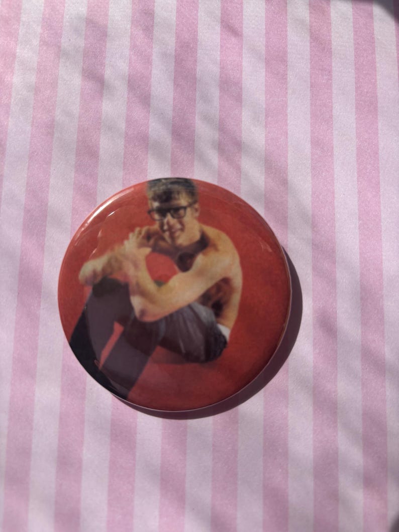 Op de afbeelding: Een ronde, rode button met een foto van een persoon met een bril die zit. De button heeft een glanzende afwerking en staat tegen een roze en witte gestreepte achtergrond.