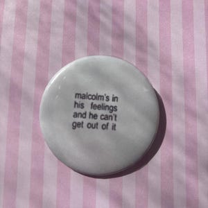 Op de afbeelding: Een witte, ronde button met de tekst "malcolm's in his feelings and he can't get out of it" in zwarte letters. De button ligt op een roze en witte gestreepte achtergrond.