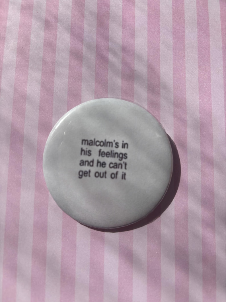 Op de afbeelding: Een witte, ronde button met de tekst "malcolm's in his feelings and he can't get out of it" in zwarte letters. De button ligt op een roze en witte gestreepte achtergrond.