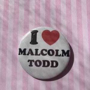 Op de afbeelding: Een ronde button met de tekst "I MALCOLM TODD" in zwarte letters en een rood hart. De button is wit met een glanzende afwerking en ligt op een roze en wit gestreepte achtergrond.