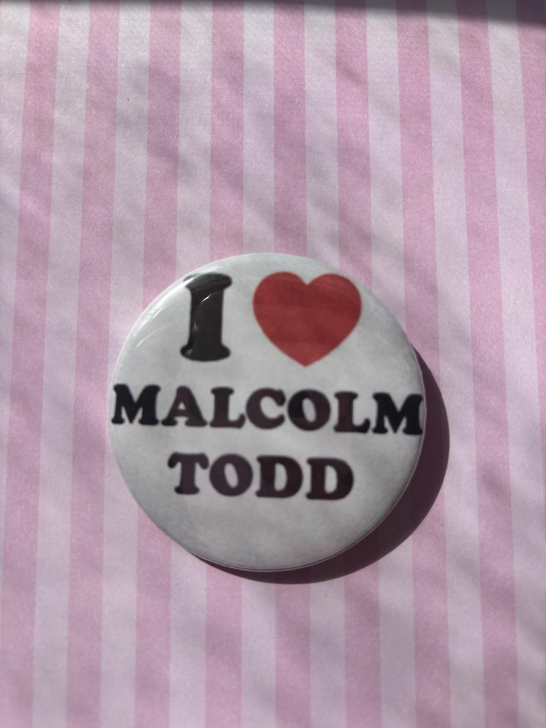 Op de afbeelding: Een ronde button met de tekst "I MALCOLM TODD" in zwarte letters en een rood hart. De button is wit met een glanzende afwerking en ligt op een roze en wit gestreepte achtergrond.