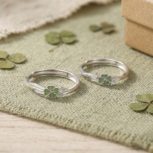 May include: Two silver rings with a green four-leaf clover design. The rings are on a green textured surface with scattered green leaves. The rings have an adjustable band.