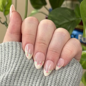 May include: Oval-shaped artificial nails with a light pink base and a white tip. The tips are decorated with a delicate floral design in pink and green. The nails are on a hand wearing a gray sweater.