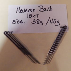 Puede incluir: Primer plano de diez agujas de barba inversa plateadas. Las agujas están dispuestas en dos grupos de cinco, con las puntas enfrentadas. Una tarjeta blanca con escritura negra es visible en el fondo. La tarjeta dice "Reverse Barb 10 CT 3ea. 389/40g".