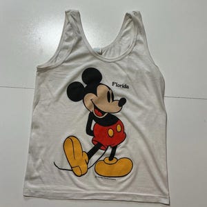 May include: White tank top featuring a large graphic of Mickey Mouse in his classic pose. Mickey is wearing red shorts and yellow shoes. The word "Florida" is printed above Mickey's head.